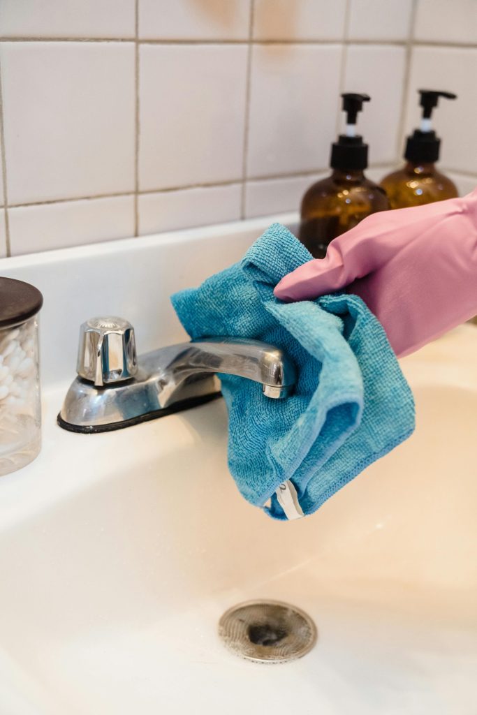 Close-up of a person sanitizing a bathroom faucet using a blue cloth and latex gloves.